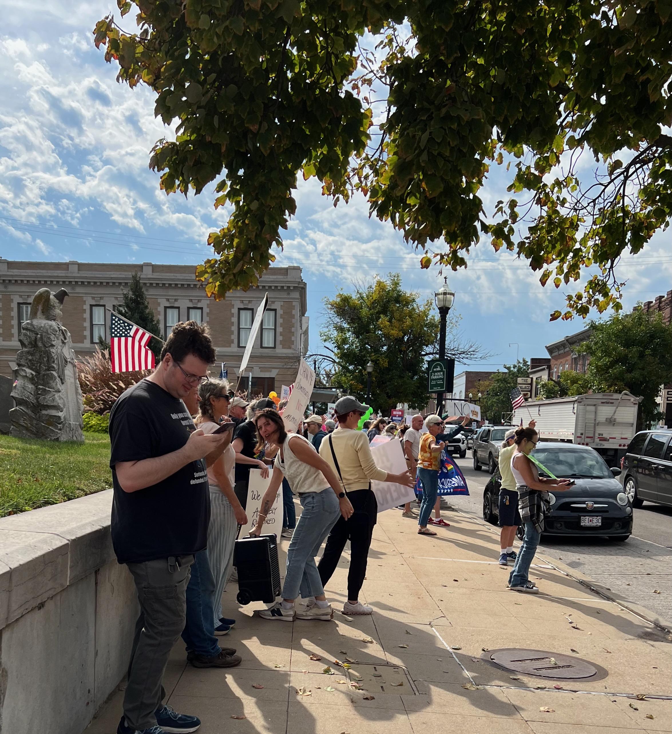 A group of about 150 protestors standing in front of a low wall along a city street with signs and banners.