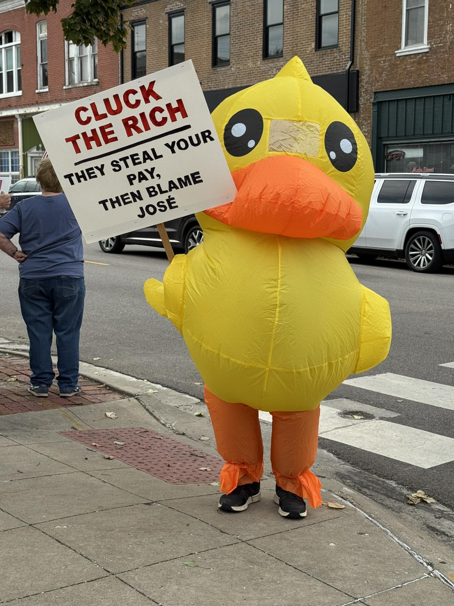 A an inflatable duck holding a sign that says "Cluck the Rich, They steal your pay then blame Jose."