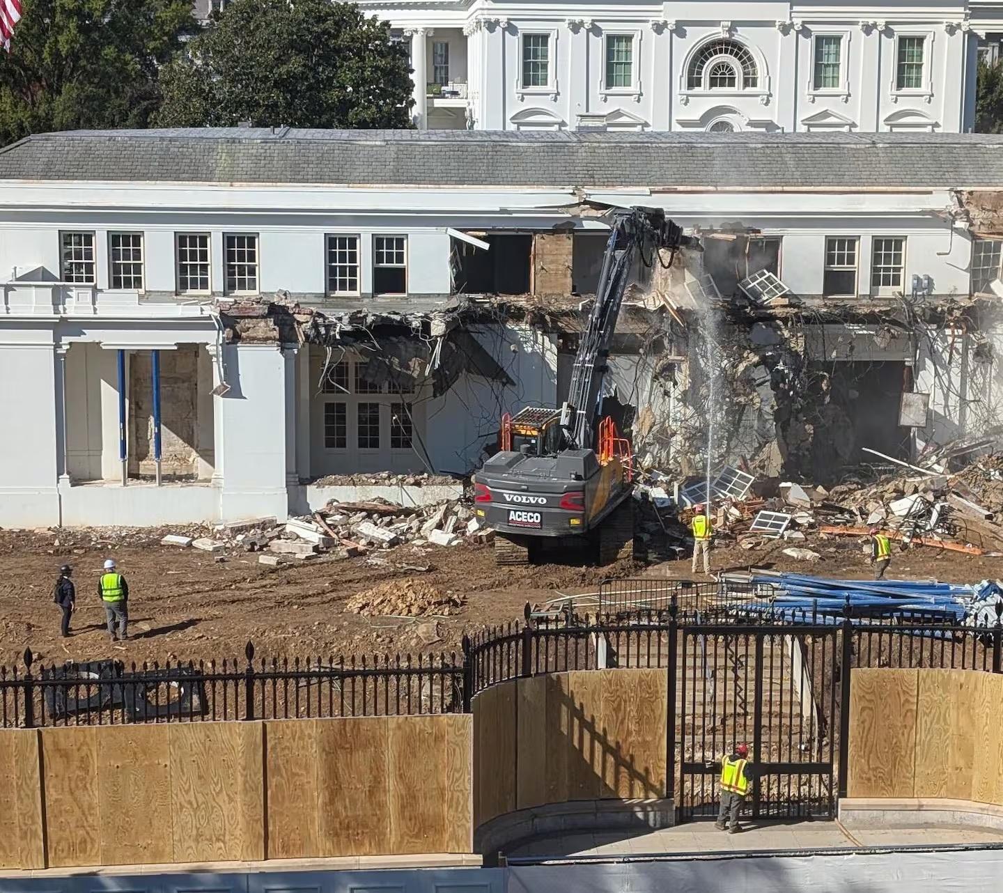 A large, classical-style building is undergoing demolition, with a significant portion of its facade removed, revealing the interior structure. A yellow excavator with “VOLVO” and “ACEDO” visible on its side is positioned in front of the building, appearing to be actively involved in the dismantling process. The building features white columns, numerous windows, and a symmetrical design, but is now partially destroyed. A black fence runs across the foreground, separating the viewer from the construction site, and several people wearing safety vests are visible in the background. The ground is covered in dirt and debris from the demolition work.