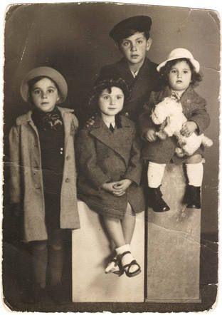 Studio photo of four siblings. Three girls (one standing and two sitting) in front, a boy in the back. They are all dresss nicely, the youngerst girl is holding a toy dog.