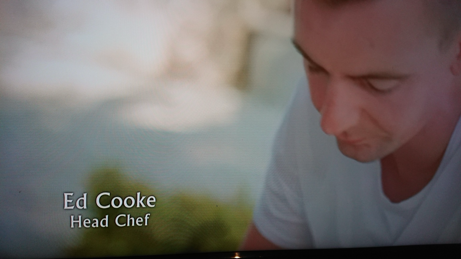Photo taken of a TV screen showing a man in a white t shirt. Next to him is text of his name and his job. He is Ed Cooke, and he is a Head Chef