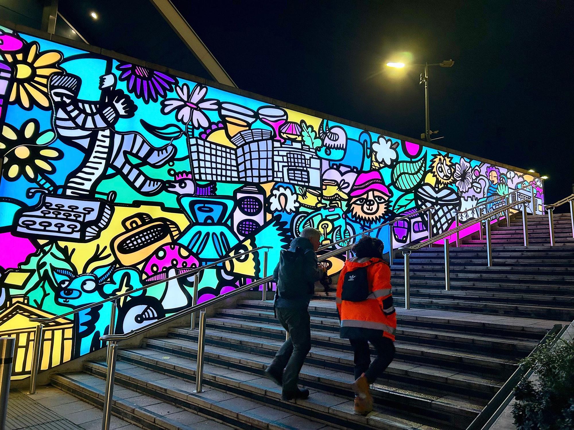 A exterior night scene with two people climbing steps beside an extra-large illuminated mural. The 51-metre long artwork outside Reading railway station is full of references to the people, landmarks, and associated creations of the town — most notably in this section, Banksy's "Create Escape" from the wall of Reading Prison and Michael Bond's Paddington Bear. "Rivers, Ruins & Regency" [2024] by Kev Mundy, in collaboration with sculptor Stuart Melrose. Station Hill, Reading, Berkshire, UK. Photo by Gearóid Burke.