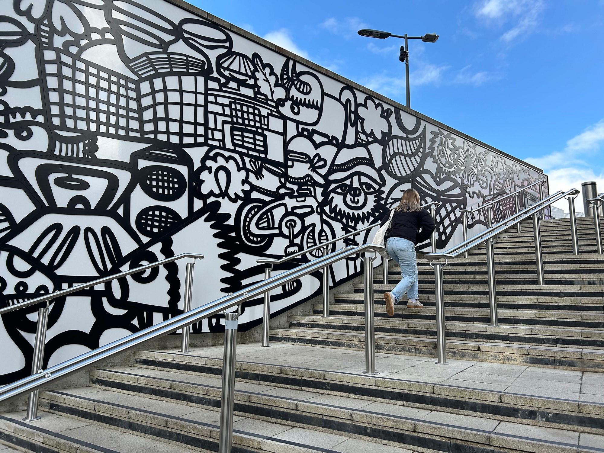 A woman climbs a series of outdoor steps beside a large figurative piece of art. The 51-metre long artwork outside the Reading railway station is full of references to the people, landmarks, and associated creations of the town — most notably in this section, Paddington Bear. Michael Bond, the author of the books, grew up in Reading and his visits to its station started a love of trains. Art by Kev Mundy, in collaboration with sculptor Stuart Melrose. Station Hill, Reading, Berkshire, UK. Photo by Gearóid Burke.