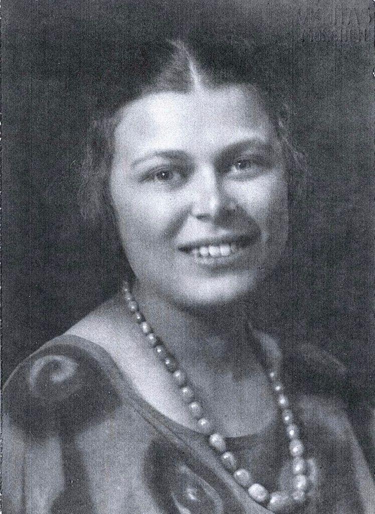Black and white portrait of a woman smiling, wearing a string of pearls and a draped blouse.