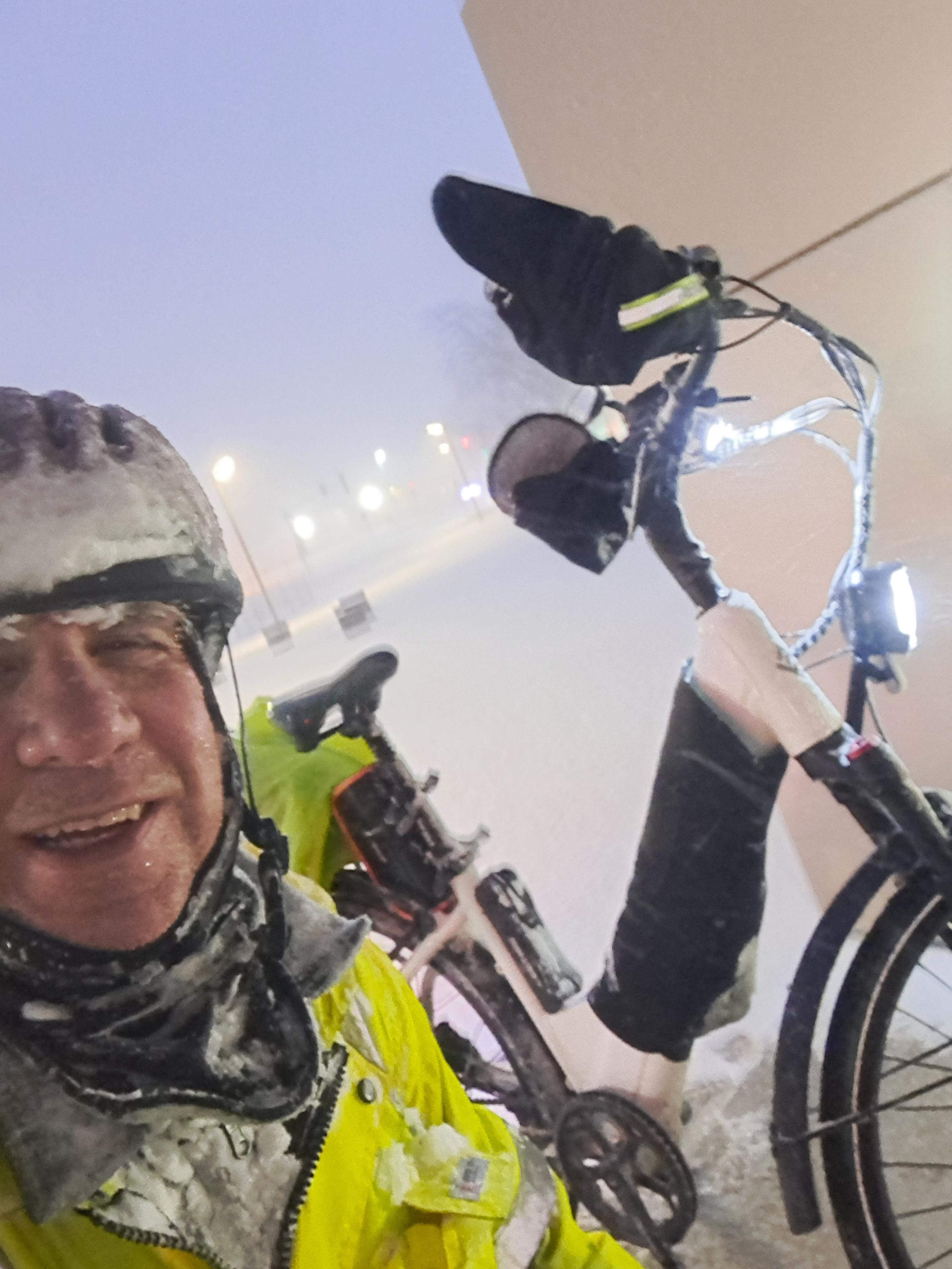 Close selfie of white man in his 50's smiling wearing a bike helmet and yellow reflective jacket kneeling next to an e bike covered in snow. He is in the protected entrance to a building.The background on the right is a tan wall the middle and left is a very grey sky with street lights in the distance. Snow is falling in big flakes giving everything a dream like appearance.