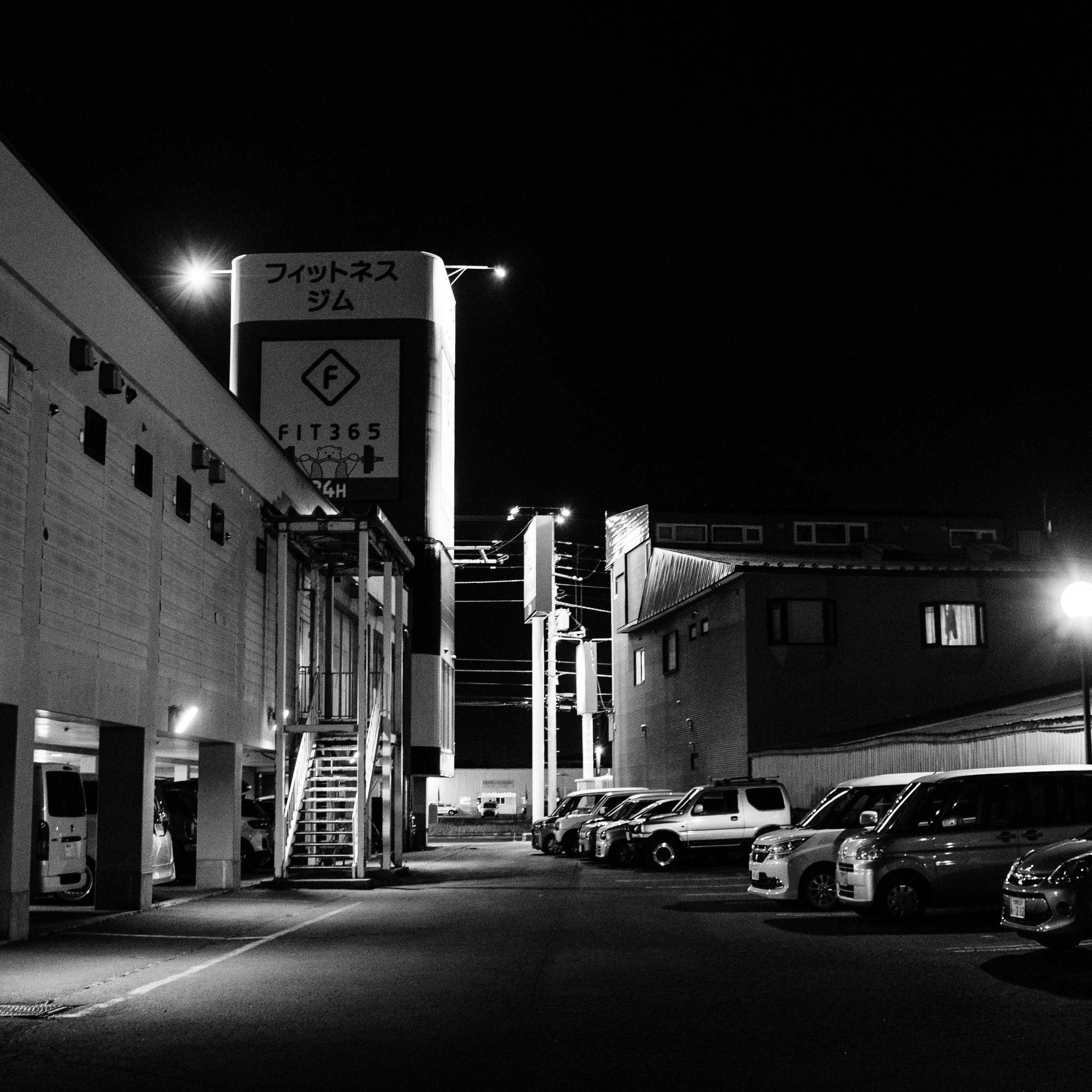 This black-and-white night photograph shows a parking lot between buildings. A large, illuminated vertical sign stands prominently. Several parked cars line the right side and an open garage area on the left. A metal staircase leads upward, and streetlights cast sharp shadows across the paved ground.