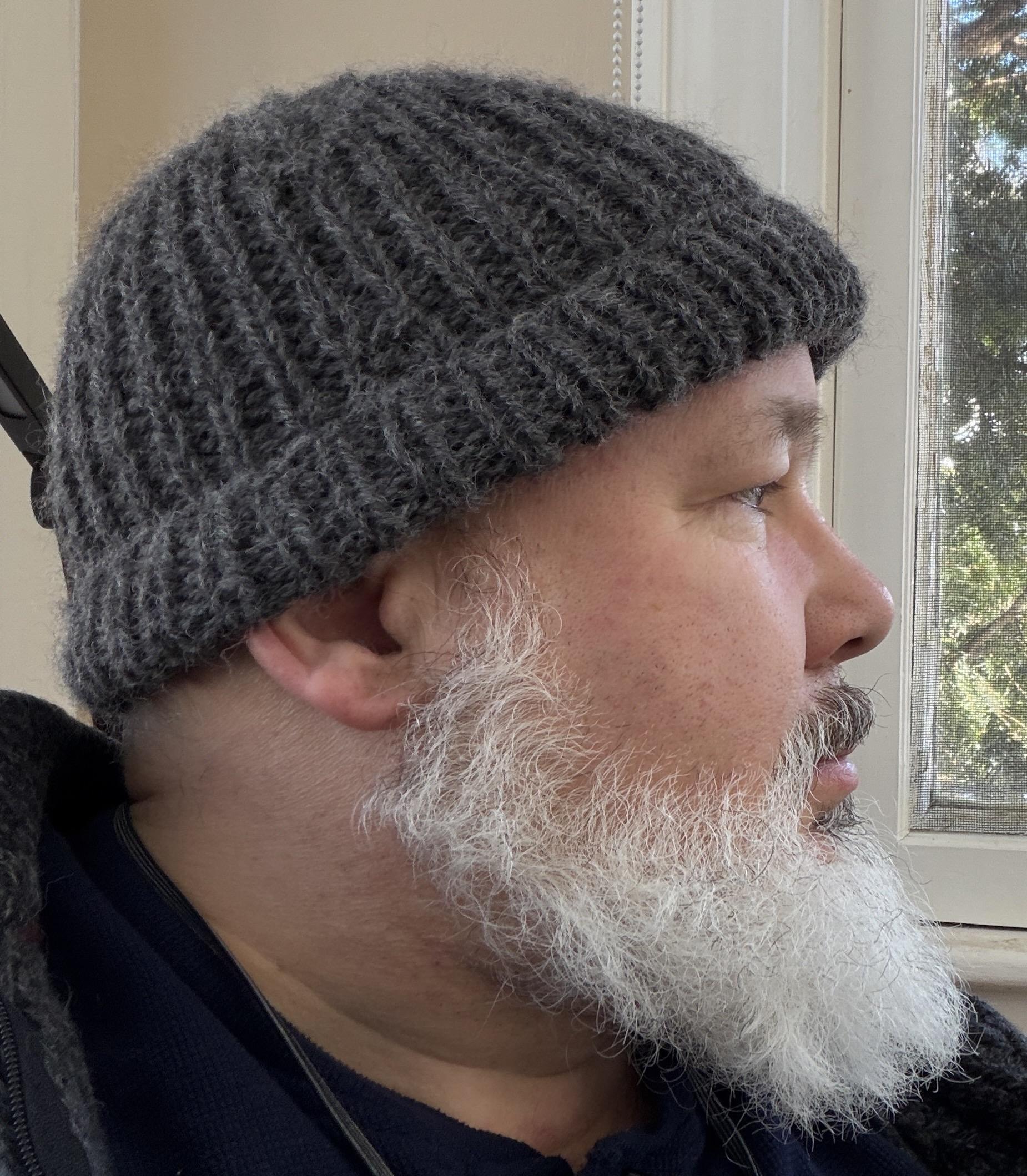 Amply bearded man in late 50s in mid-dark gray rolled brim knit cap that covers his luxuriously bald head. He is far prouder of this self-knit accomplishment than is deserved.