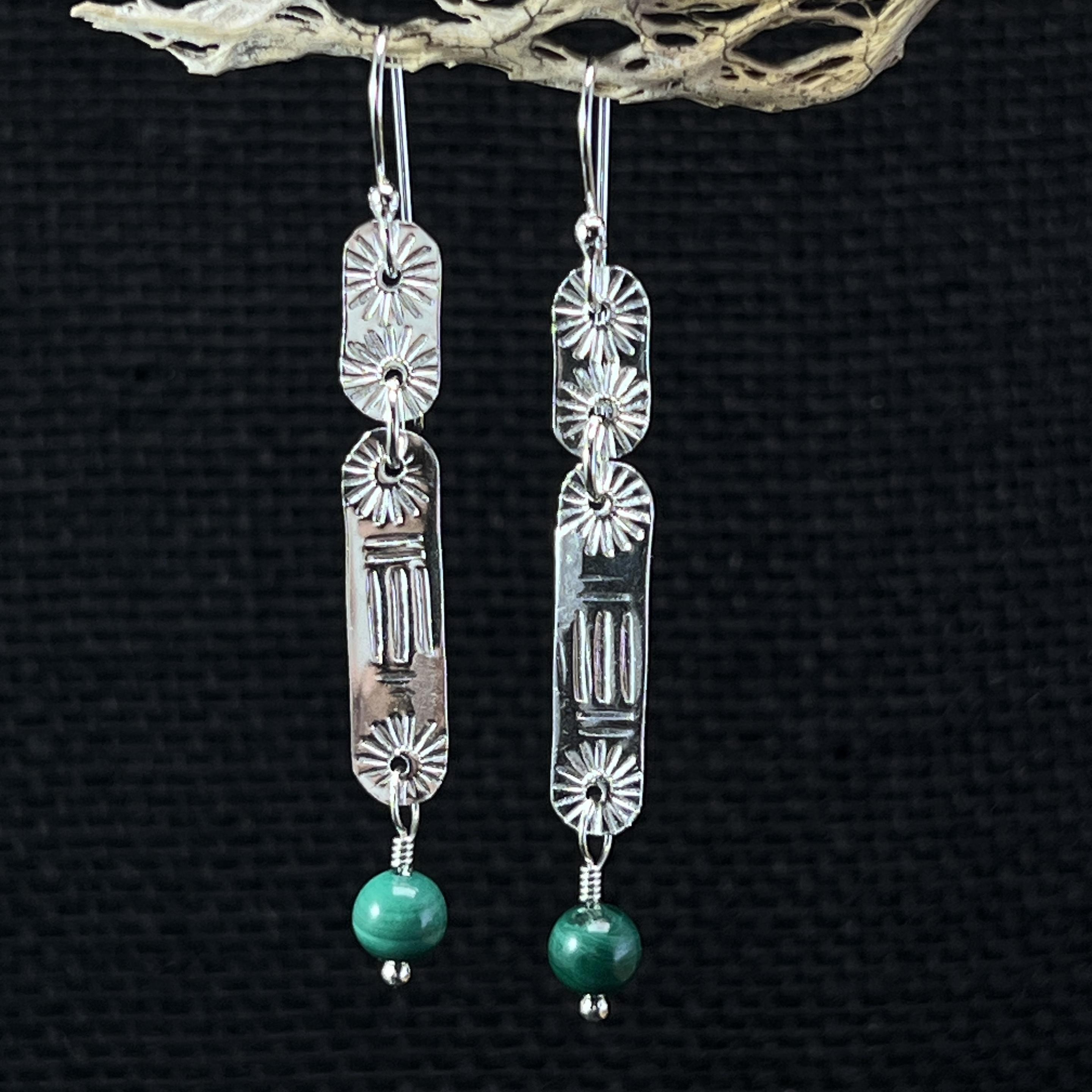 Photograph of a pair of sterling silver earrings with Malachi beads. Each earring is made from two strips of silver, one that is a half inch and the other that is a full inch long, that have been stamped for a pattern or texture. The pieces are joined together with silver jump rings. The beads are attached at the bottom with a wrapped loop. The ear wires are sterling silver. The copper version of these earrings will be identical except made with copper strip and wire and a black niobium ear wire that includes a copper bead.