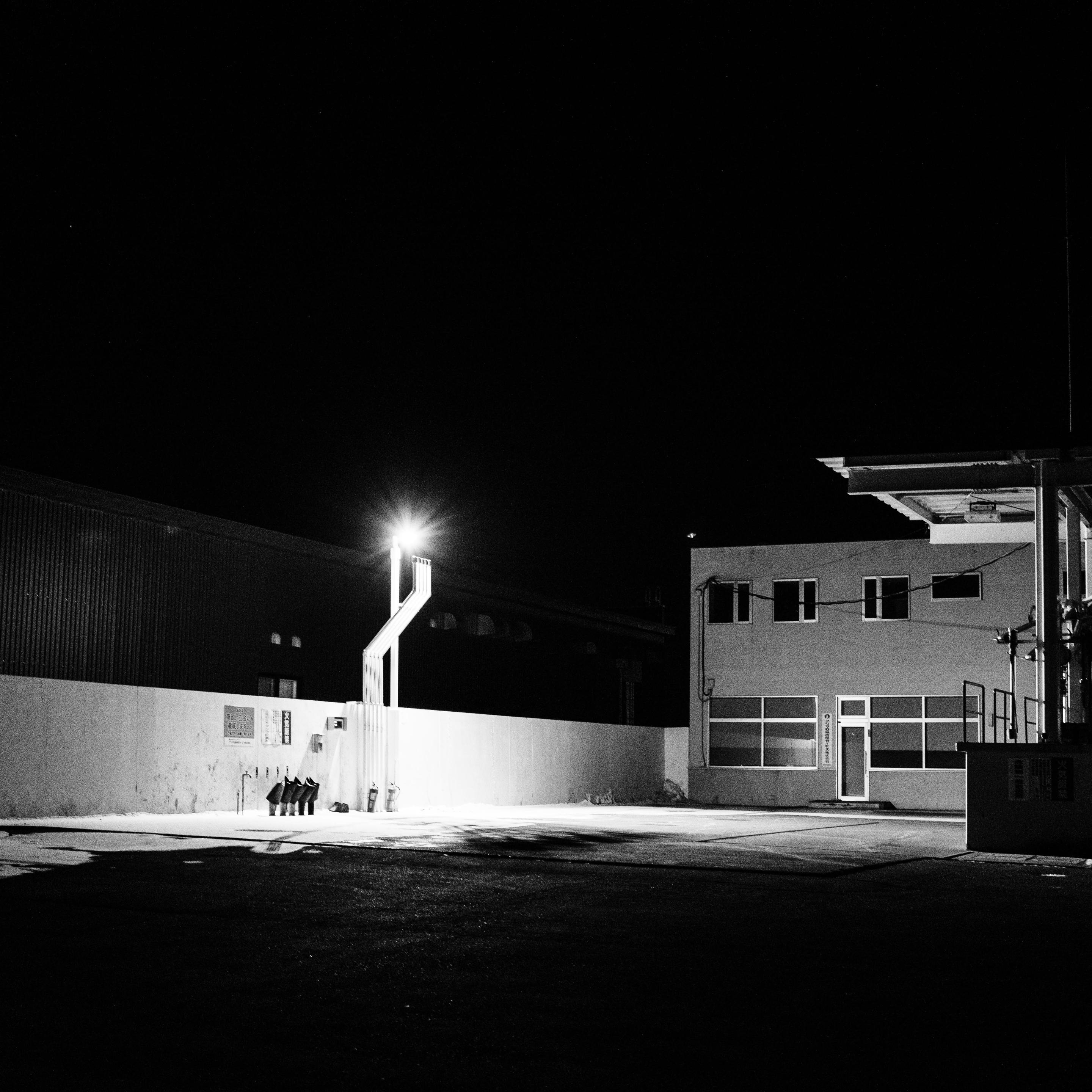 This black-and-white night photograph shows an industrial area. A bright light source casts a starburst pattern, illuminating a long white wall and several vertical pipes. To the right stands a two-story building with windows and a door. The dark foreground is paved, while the background sky is entirely black.