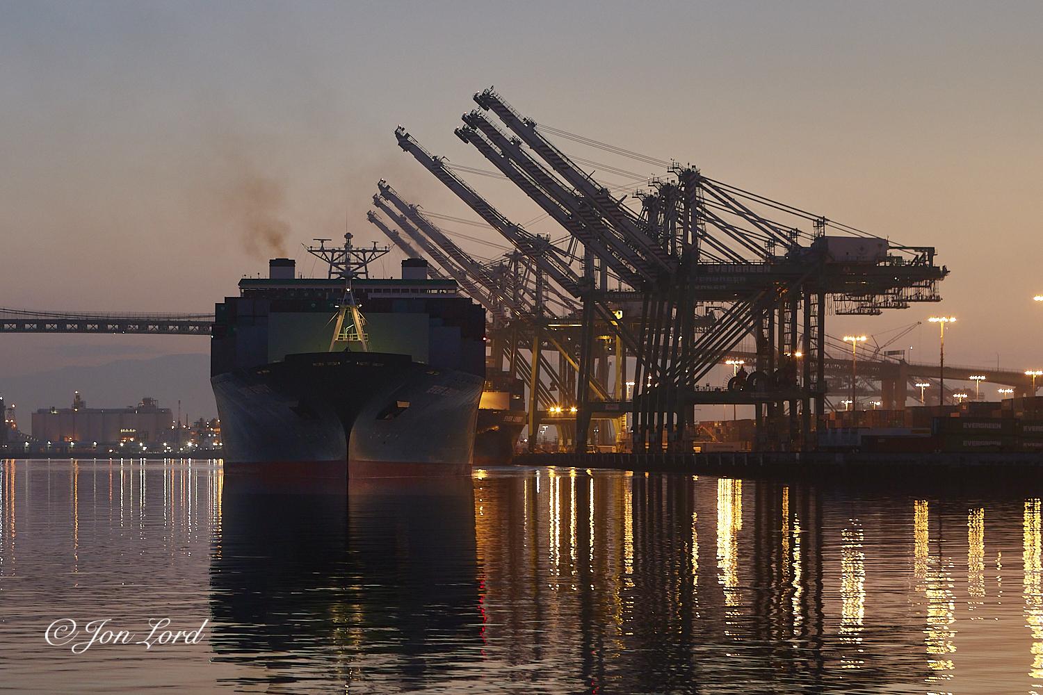 This is a colour dawn photo in landscape format of a large container ship leaving port. Port of Los Angeles, California (2018).

The lower third of the photo is filled with dead calm harbour water with just the smallest ripples disturbing the surface. The water on the right side has vivid and colourful reflections from the port's floodlighting. On the right side is a row of 7 gantry cranes, used for the loading and unloading of shipping containers, all with their booms raised. Behind them and further to the right is the container terminal with rows of stacked containers. To the left of centre is the front of a large container ship heading towards the camera at dead slow, so slow the vessel is not creating any wake. Only the front and some of the top surfaces are in view. 2 or more metres of red anti-fouling can be seen above the waterline. The hull, while in shadow, is a dark green. There is a small floodlit mast above the bow. Also in shadow are the thousands of stacked shipping containers above the deck. Out of view, there are many stowed below deck. Above the upper row of containers the bridge can be seen with a long line of rectangular windows stretching across the width of the ship. Above and behind the wheelhouse are two funnels, one on either side. The starboard funnel emitting a trail of brown smoke while the port funnel has a heat haze above it. In the background is a warm orange glow from the still to rise morning sun.