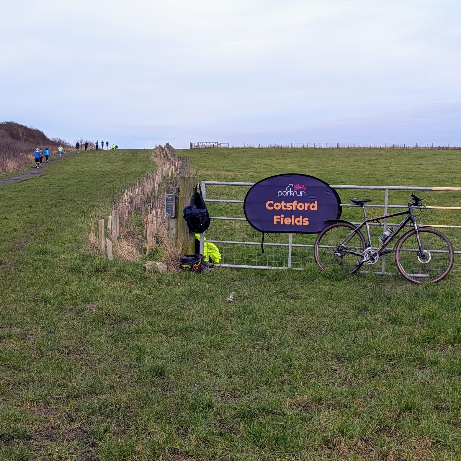 A photograph of the Cotsford Fields parkrun sign on a metal gate with a bike beside it. A group of runners are running up a hill in the distance.