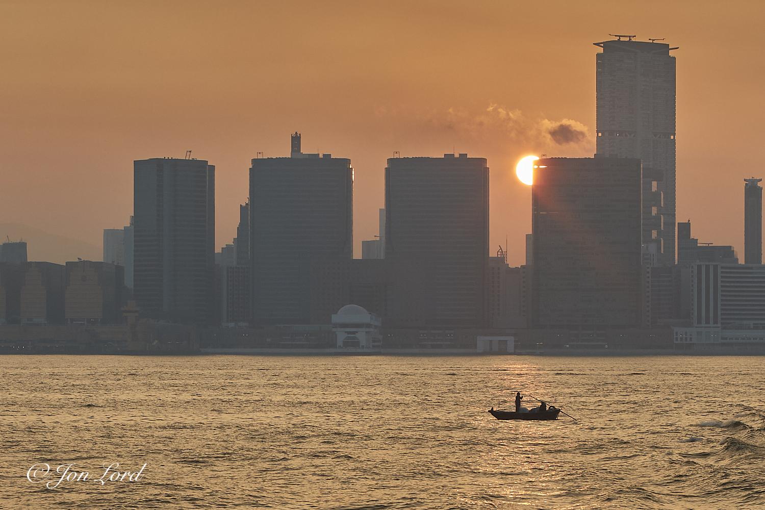 This is a colour sunrise photo in landscape format of two people fishing from a small dinghy with the sun rising between a city high-rise in the background. Victoria Harbour, Hong Kong (2017).

The deserted harbour water rises upwards one third in the image. The water is slightly disturbed with small wavelets breaking the surface. To the lower right side of centre and perhaps about 100m away is a small dinghy (skiff), no more than 3m long and deep in shadow. On the dingy is a person standing in the centre with another sitting in the stern with a small pile of possibly nets. Attached to the transom (the flat stern of the boat) is a long oar that stretches to the middle of the dinghy and that's used to propel and steer. In the background and about 1km away is a waterfront that stretches from right to left. Behind is a high-rise cityscape with the still low morning sun, partially obscured, rising behind and to one side of one of the skyscrapers. Above is a clear and nearly cloudless sky rendered a deep orange by the early dawn light.