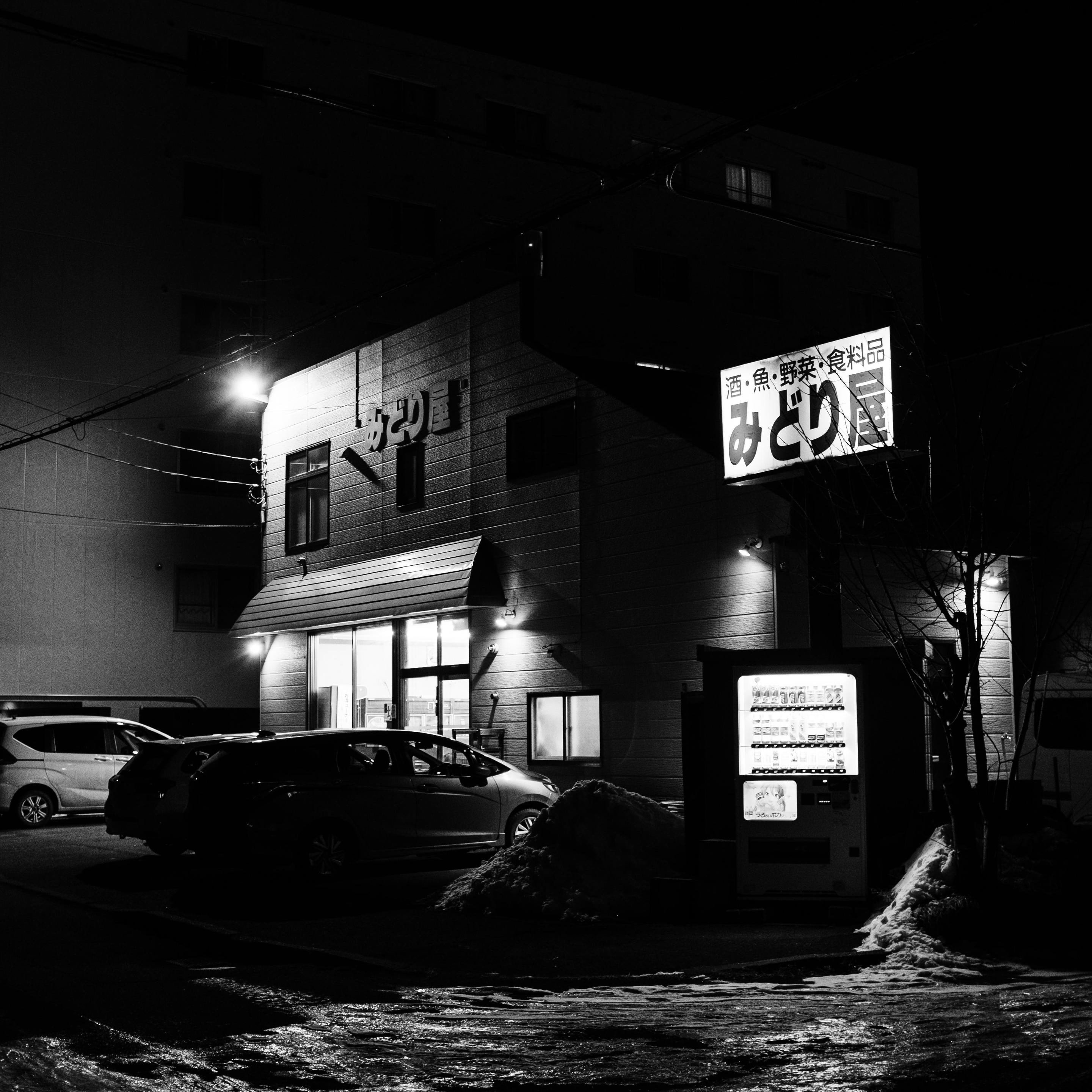 This black-and-white night photograph shows a two-story commercial building with a brightly lit entrance. Several cars are parked in a lot in front. To the right, an illuminated vending machine stands near snow piles. A large dark residential building and power lines are visible in the background.
