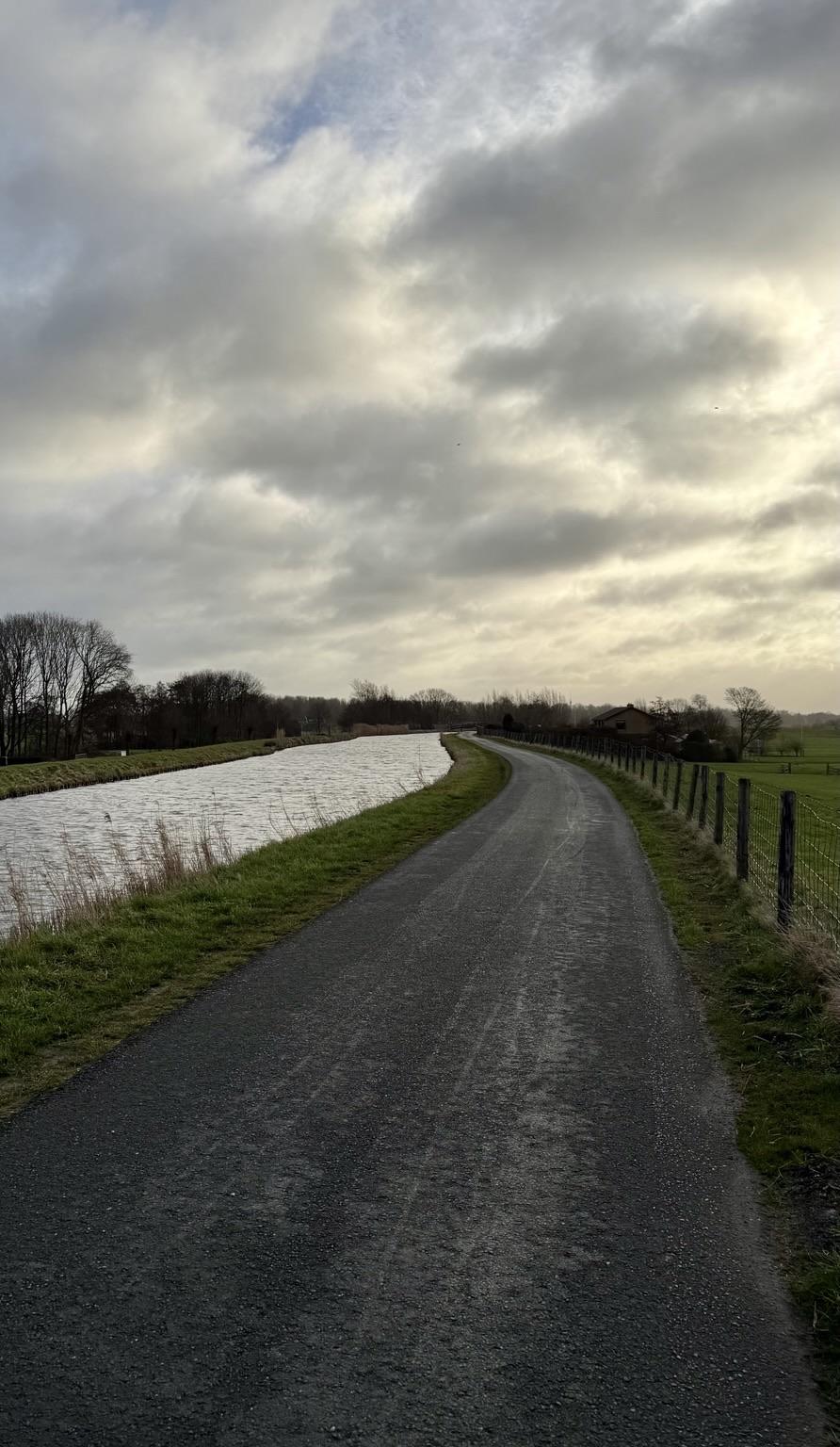 Een fietspad naast een brede polder sloot links met daar achter weilanden omzoomd met bomen en struiken, rechts weilanden.De lucht is bewolkt met verspreide wolken en bomen zijn zichtbaar op de achtergrond.