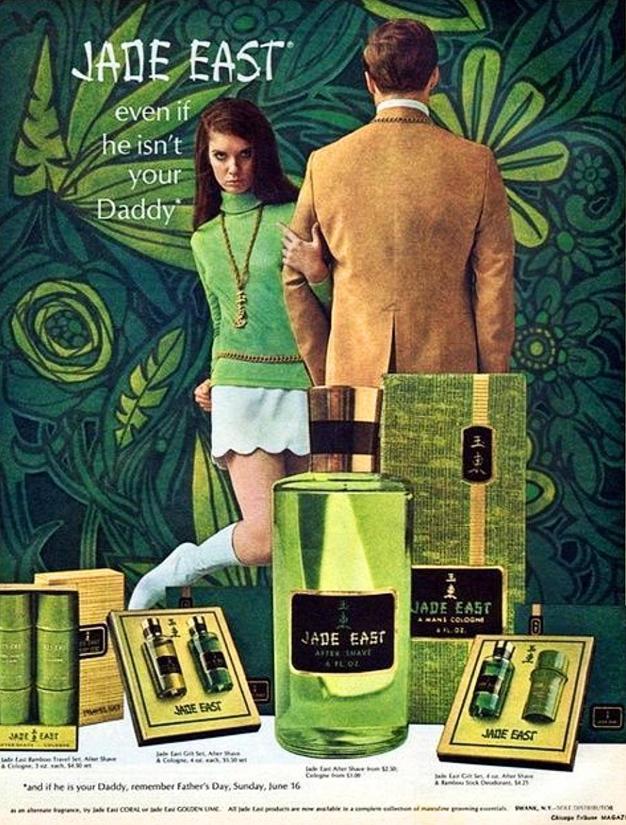 A late-1960s magazine advertisement for Jade East men’s cologne features a stylised, mod tableau set against a dense, psychedelic green backdrop of swirling botanical motifs. In the foreground, a large, translucent green bottle labelled “Jade East After Shave” is flanked by boxed gift sets and smaller product displays. Behind the products stands a sharply dressed man in a tan jacket, shown from the back, while a young woman in a bright green top, pale mini skirt, and knee-high white boots clasps his arm and gazes directly at the viewer with a cool, provocative intensity. Bold white lettering at the top reads “JADE EAST — even if he isn’t your Daddy,” underscoring the era’s playful yet suggestive marketing tone.