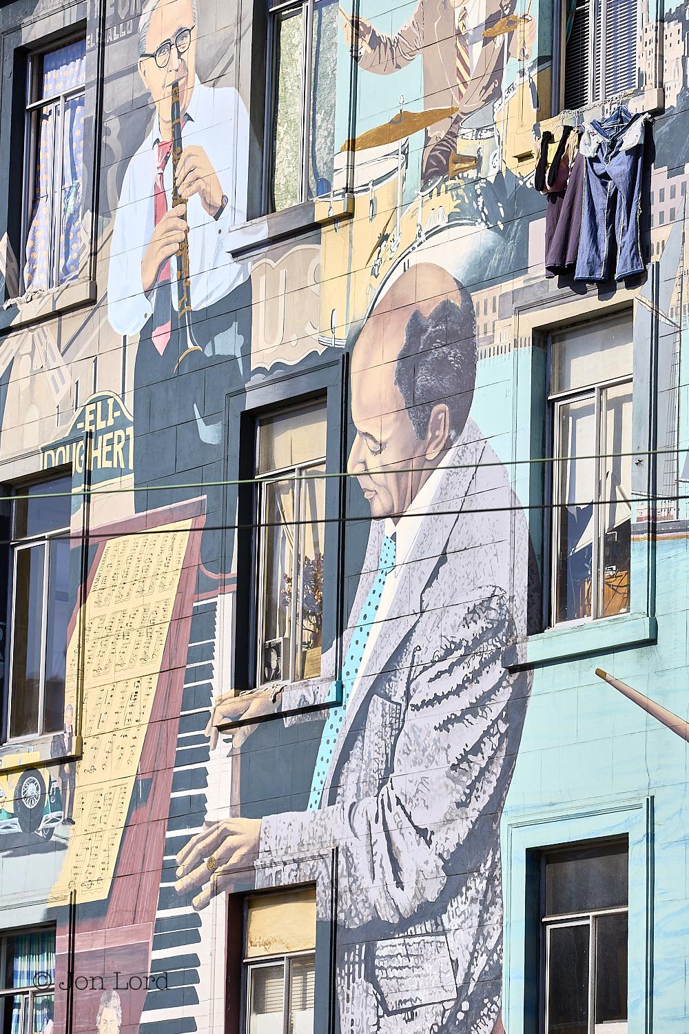 This is a colour street photo in portrait format of the front of a building brightly painted with the figures of various famous jazz musicians playing their instruments on a back drop of local street scenes. Columbus & Broadway, San Francisco (2013).

The facade of the building is viewed obliquely from an angle of about 45º and captures 3 floors above the ground level. The building is a city low-rise apartment block that's neither new nor old. In view are the three floors with three windows on each floor, some open, some closed. Some with curtains drawn, some open. One with laundry drying below the window cill. Stretching upwards from the base to a little above the centre of the photo and dominating the image is the mural of a middle-aged pianist dressed in a jacket and blue tie,  playing the piano with sheet music laid out above the keyboard. Above the pianist and in the upper left corner is a further middle-aged man wearing glasses, shirt and tie and playing the clarinet, Benny Goodman perhaps ? To his right and in the upper right corner is a man playing the drums, his head out of view. The background of the facade in the lower left corner is light blue with the gaps between the musicians being filled with local scenes with street signs, a windmill, sailing boat, car and high-rise in view. 

This is one of the bright and colourful murals that decorate in and around San Francisco's Chinatown.