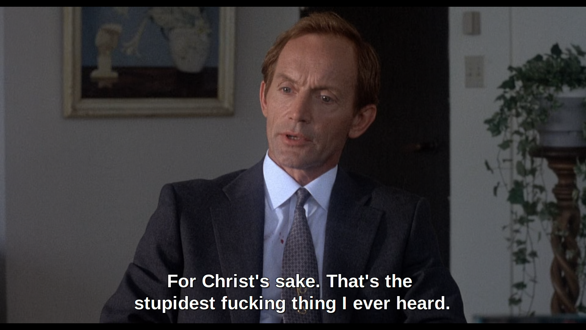 Frameshot of Lance Henriksen from the movie "The Horror Show". He's sitting inside a room and wearing a grey suit, white shirt and grey tie. He's looking to the ground and his mouth is partially opened uttering the captioned words "For Christ's sake. That's the stupidest fucking thing I ever heard."
