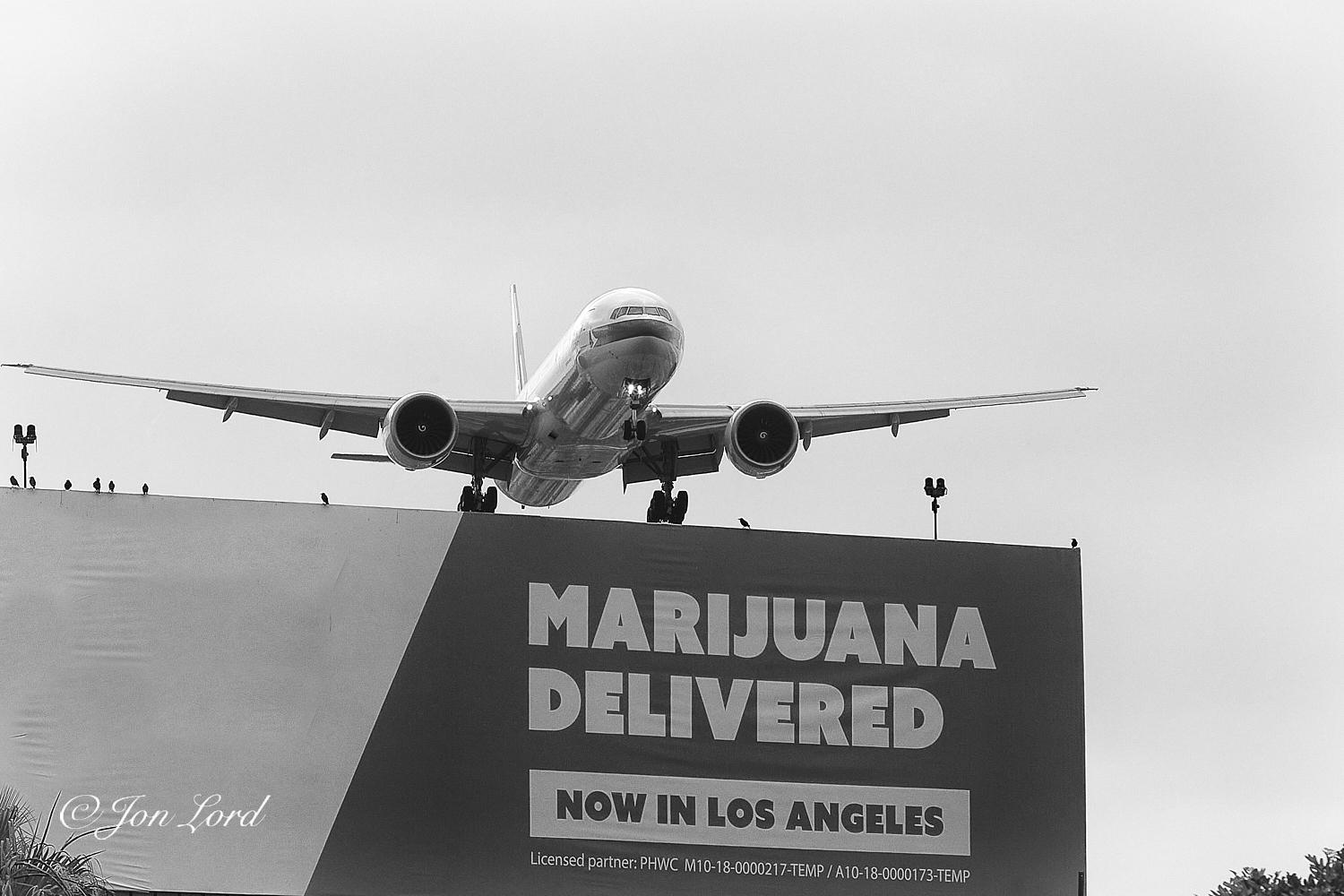 This is a black and white photo in landscape format of a landing plane flying over large billboard. Los Angeles, California (2018).

Filling the lower left quarter and stretching across to close to the right edge of the photo is a large billboard (advertising sign) above and to the side of the street with the words in bold white letters against a dark background - 
'Marijuana Delivered' 
And below in reverse video - 
'Now In Los Angeles'
Overhead the billboard is a large jet plane (Cathay Pacific Boeing 777) flying towards the camera and about to land, only a few hundred feet above the ground: it's undercarriage just caressing the top of the billboard. To the right of the sign and above the jet is an overcast sky, devoid of and detail. 