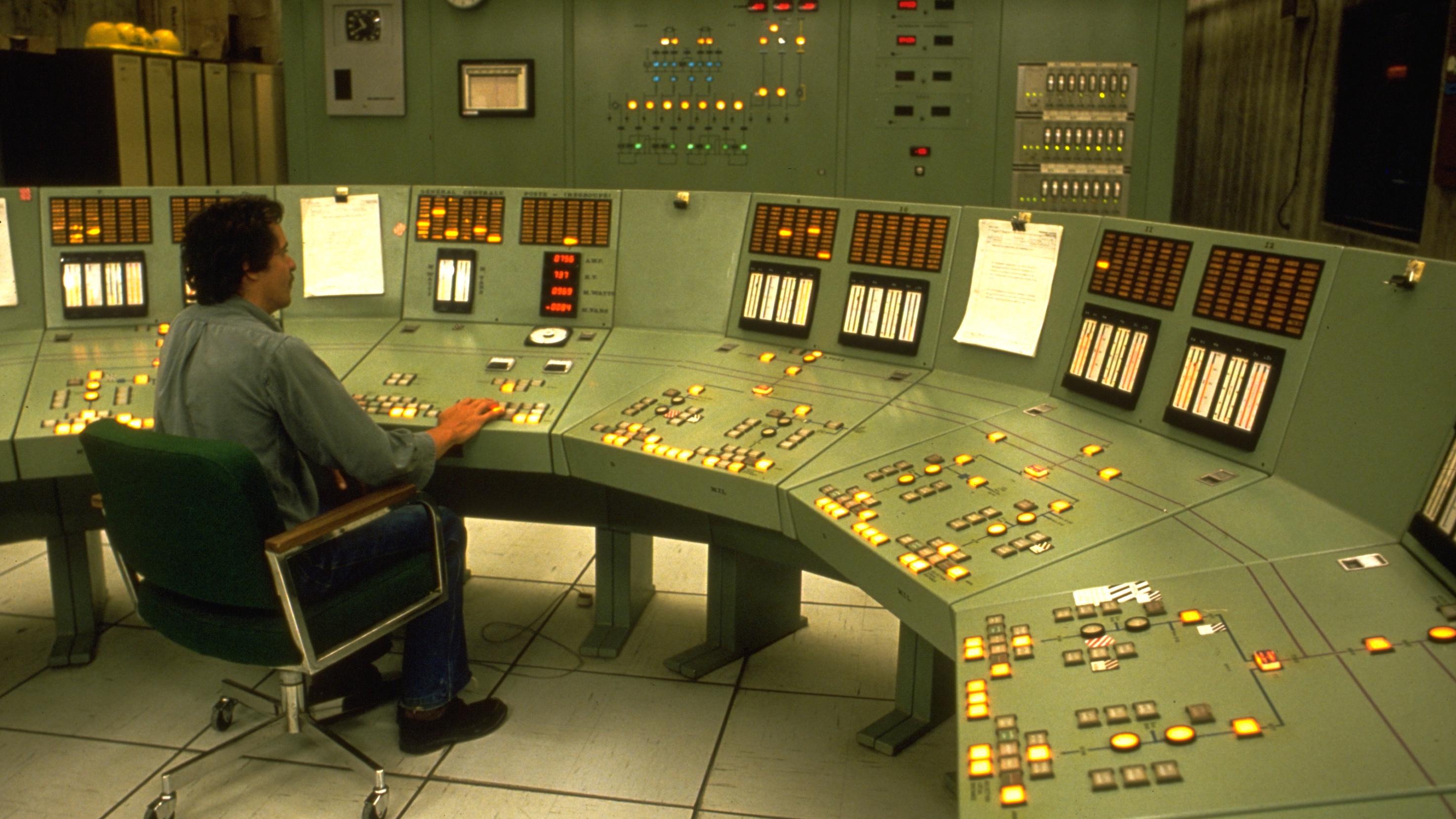 Retro stock photo of a big industrial control room with sea foam green walls and consoles. A man sits in a chair and manipulates the controls.