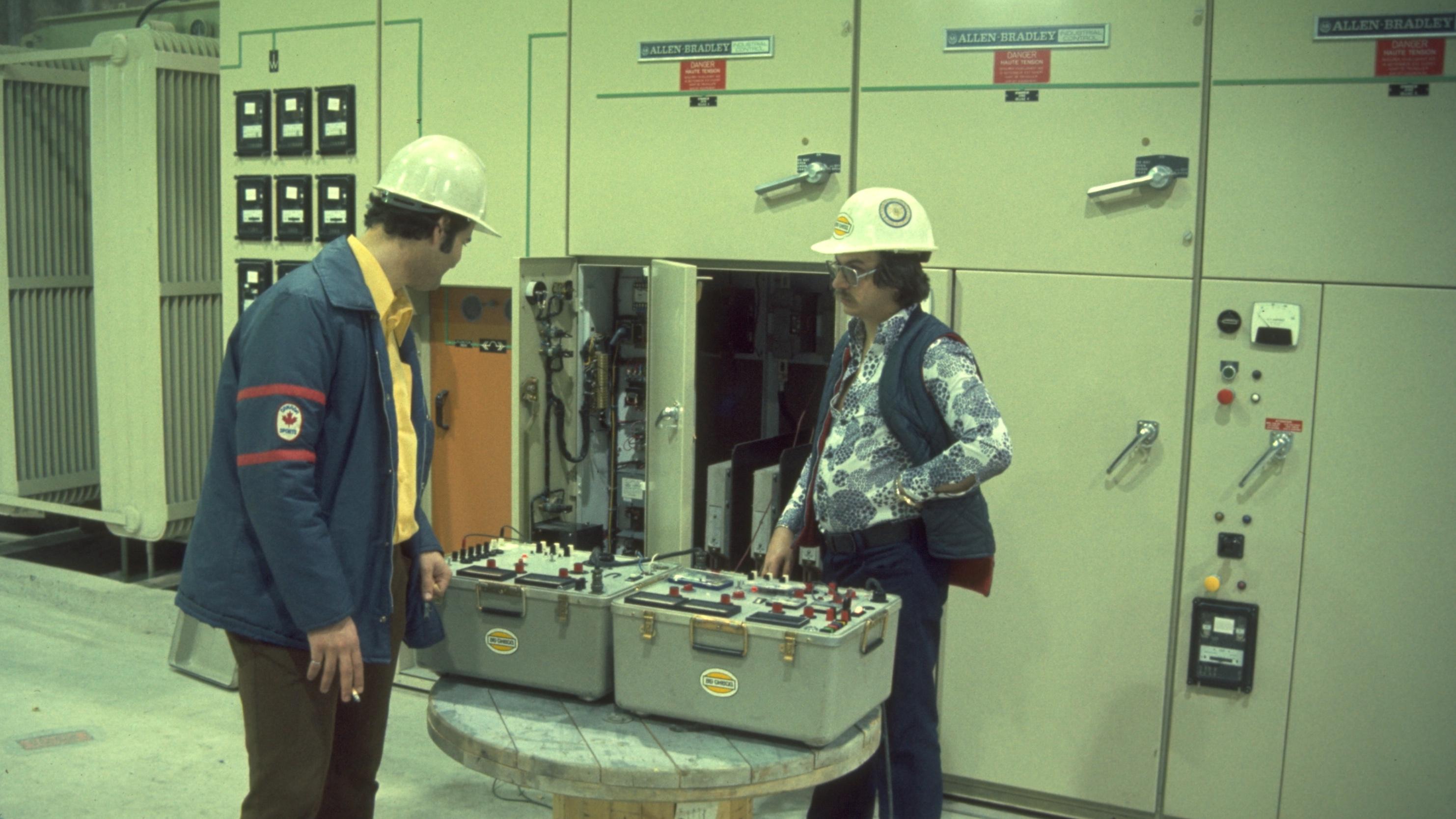 Retro stock photo of a big industrial room with light sea foam green walls and machinery. Two mean wearing hardhats examine large modular industrial control boxes on a wooden table. One of the men is wearing a very floral print shirt with wide lapels.