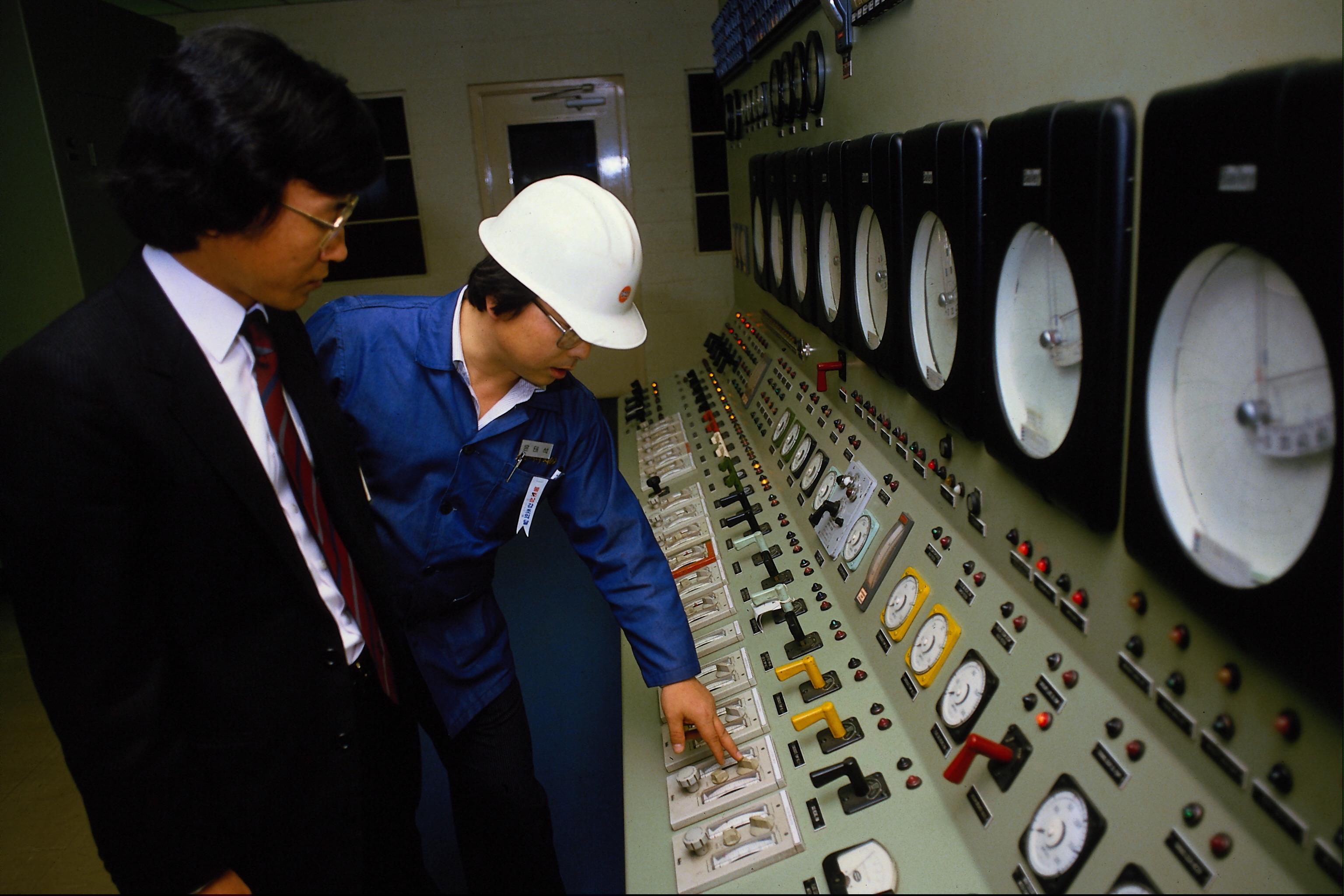 Retro stock photo of an industrial control room with sea foam green walls and consoles. Two Japanese men examine the console controls. One is wearing a business suit and the other a hard hard and industrial jumpsuit.