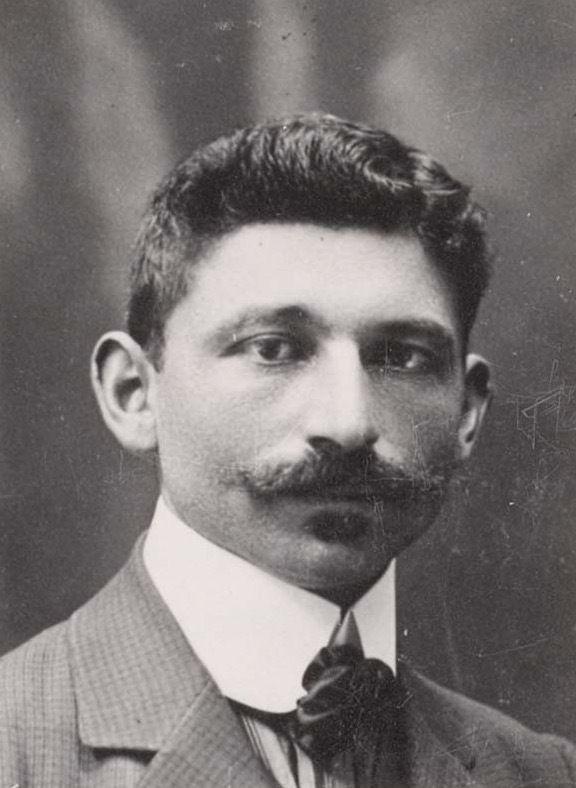 A portrait photo of the face and shoulders of a man. He has nicely styled short hair - with a curl and a mustache. He is waring a suit