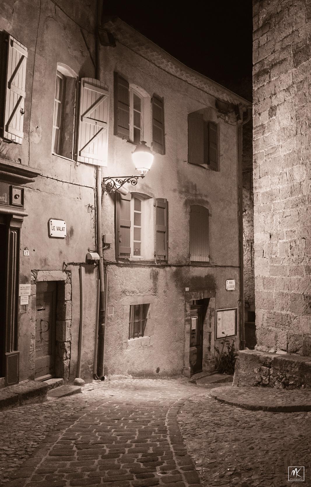 Sepia-toned photo of a cobblestone street in an old town in Provence, with a bright street light mounted to a building illuminating the scene.