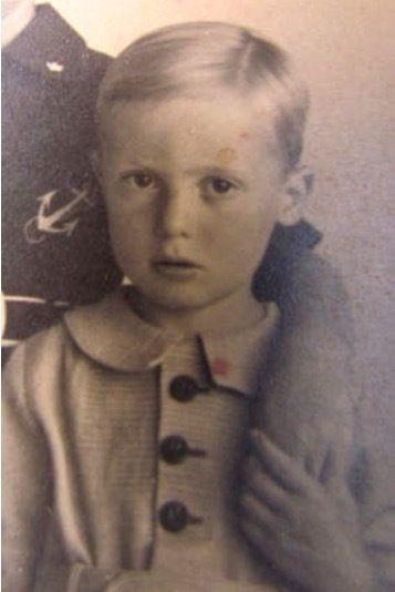 A young boy wearing a shirt with big buttons. He has short hair pulled back with a parting. Someone standing next to him embraces him with a hand on his shoulder - and the boy holds that person's hand.