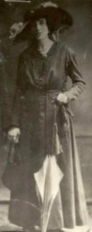 Vintage photograph of a woman standing, dressed in early 20th-century attire with a large hat and holding a handbag.