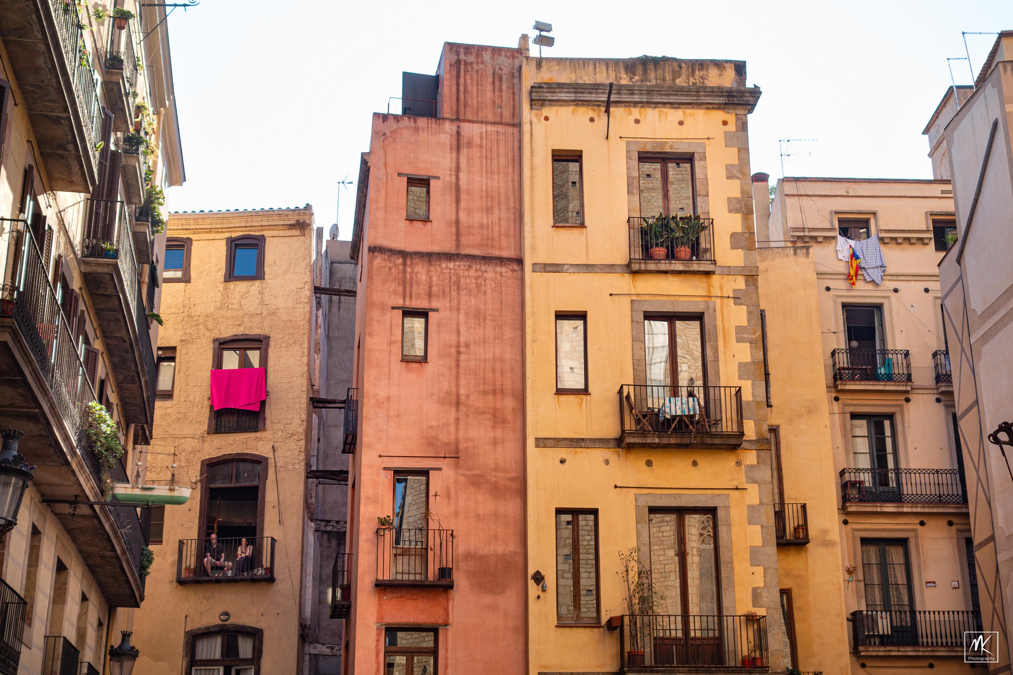 Color photo of yellow and orange buildings above a plaza in Barcelona with two people sitting and looking out from a small balcony on one of the buildings.
