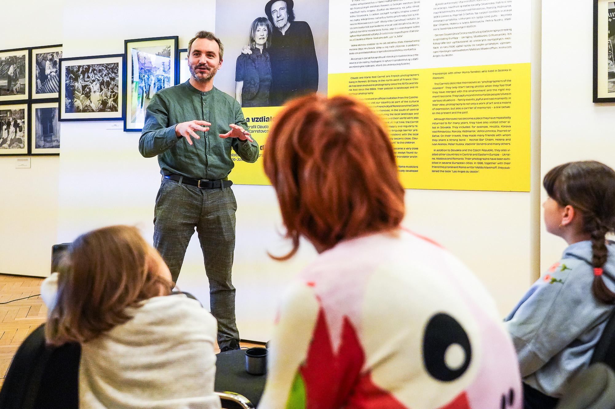 A man speaks to an audience inside the exhibition space, gesturing with his hands while seated listeners face him; behind him are photographs and a large portrait display.