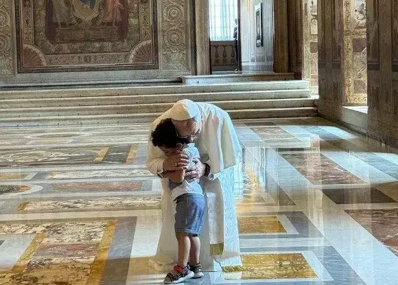 An adult in white robes is embracing a child in an ornate indoor setting. The child is wearing a white t-shirt, blue shorts, and grey sneakers. The floor features large, multi-colored marble tiles in patterns of white, tan, tan, and grey. In the background, there are stone steps and walls decorated with classical-style paintings.