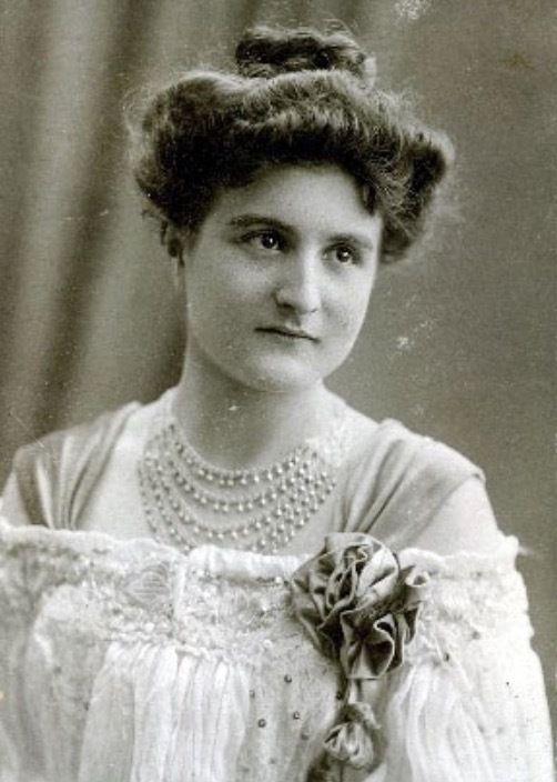 Vintage black-and-white portrait of a woman dressed in an elegant, early 20th-century outfit with detailed embellishments and multiple strands of pearls.