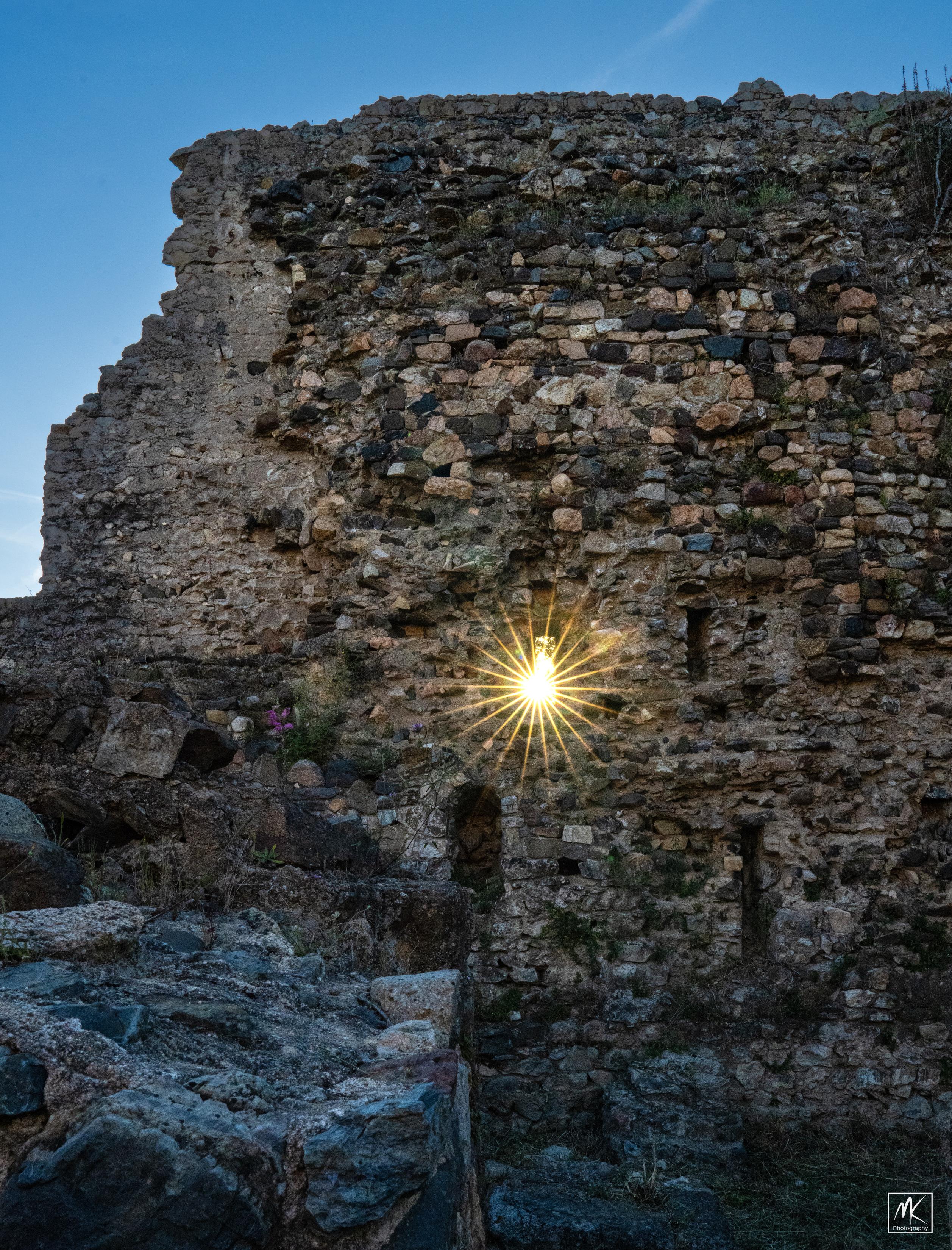Color photo of a rayed burst of light from the setting sun passing through a narrow vertical window or loop hole in the ruined stone wall of a medieval castle.