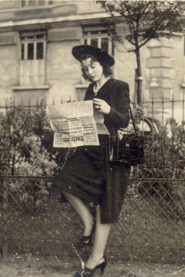 A woman in vintage clothing stands outdoors reading a newspaper. The attire includes a hat, jacket, knee-length skirt, and heels. The background shows trees and a building.