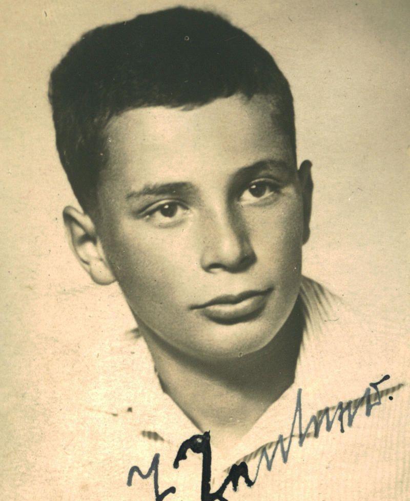 Close-up portrait of a young boy, looking directly at the camera with a serious expression. His signature is below the image.