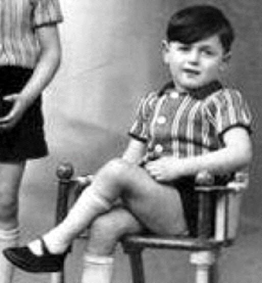 A black and white photo showing a young boy  sitting on a chair, wearing a striped shirt and shorts.