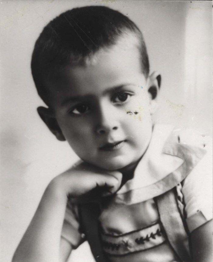 Black and white photo of a young boy with short hair, resting their chin on their left hand. The child is dressed in a collared shirt and overalls, and is looking directly at the camera.