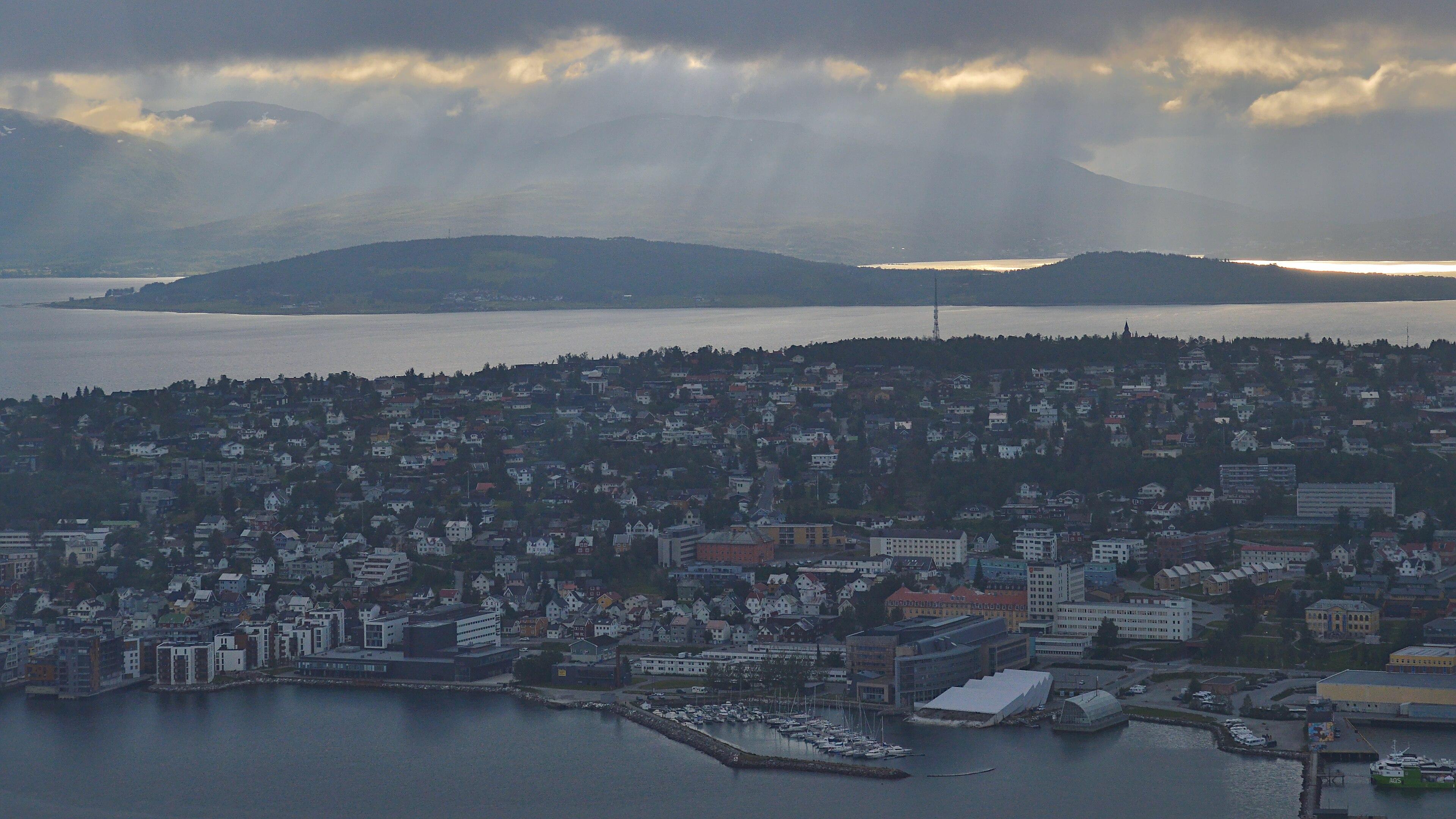 A photo looking down on the city of Tromsø. The sky is filled with clouds making the city dark. There are crepuscular rays coming through the clouds on the mountains in the distance.