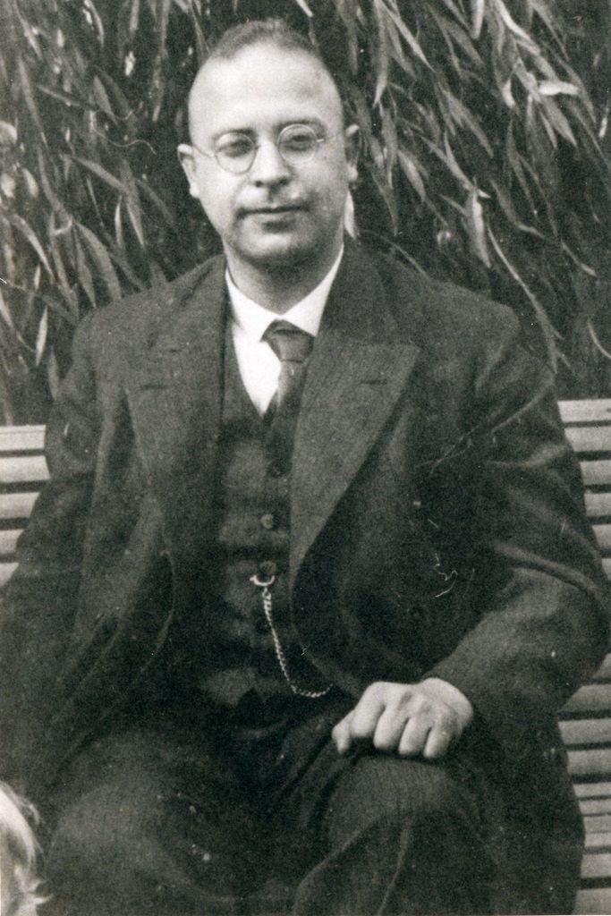 Historical black and white photo of a man seated on a bench, wearing a formal three-piece suit and round glasses. He is looking directly at the camera, with a foliage background.