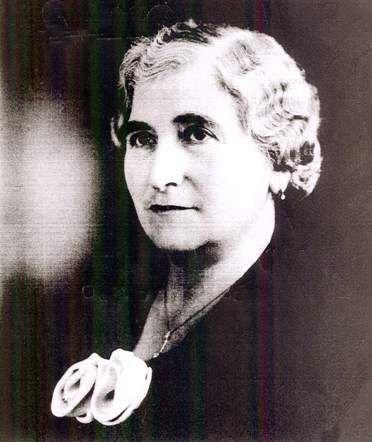 Black and white portrait of a woman, featuring a prominent white flower pinned on her left shoulder.