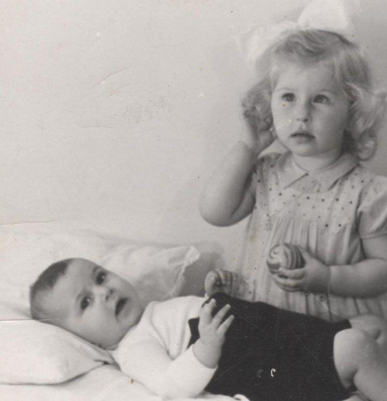 A black and white photo of a toddler sitting next to a baby who is lying down, both looking curiously towards something out of frame.