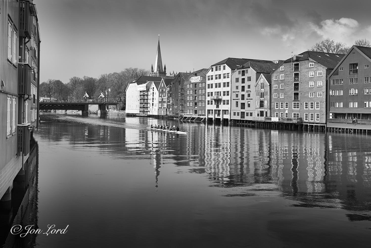 This is a black and white photo taken along an urban length of river lined with historic wooden warehouses and with a Coxed Eight competition rowing boat gliding down the river. Trondheim, Norway (2024).

Extending away from the camera along the left edge of the photo and viewed along the length of the facade is a row of old, three of four floor, wooden buildings, now apartments, that line the waterfront and curve away to the left and out of sight after about ten metres. On the opposite bank is a long line of twelve old wooden warehouses, all individual and built with the sides touching each other. The warehouses, all restored and repurposed as apartments and businesses, are view obliquely with the warehouse on the right edge of the photo being closest to the camera and with the buildings becoming more distant as they pass through the centre and to the left. The warehouses sit on a mass of wooden piles driven into the river and have five or six floors topped with large pitched roofs. Beyond the last warehouse is a line of oak-like trees with a tall, simple church spire behind. The warehouses and church spire are reflected in the slightly rippled surface of the river. In the centre of the image is our Coxed Eight (I think?) rowing boat, underway in the middle of the river and heading towards the camera. The boat is viewed obliquely with the bow and right side in view at a distance of about ten metres and is passing through the reflection of the church steeple. The boat is long, very thin and is only about half a metre above the waterline. Each of the rowers face to the rear of the boat with each rower having one long oar on alternating sides (first rower- left oar, next rower-right oar and so on) and in the stern is the cox, who happily is facing forwards and who steers the boat. In the background is an old pedestrian, steel truss bridge with two masonry pillars on the far (right) bank and with an arch over each pillar.