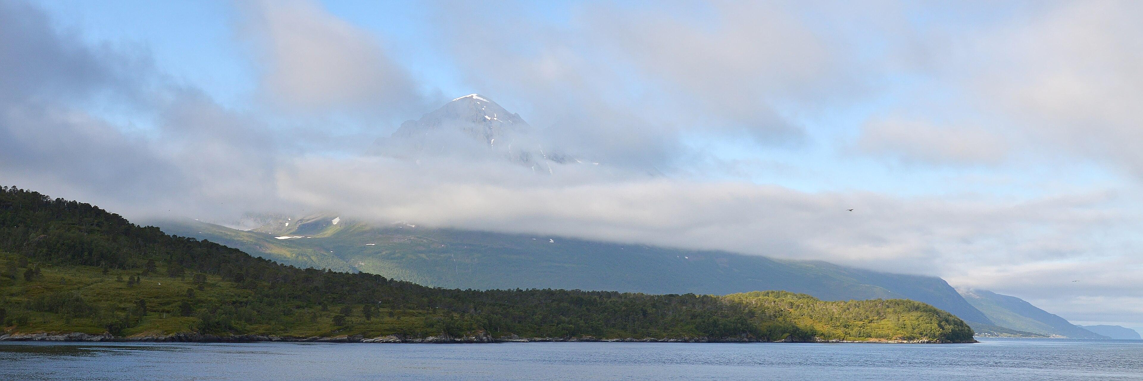 A panoramic photo of mountains leading into water. There are clouds partly obscuring the mountains.