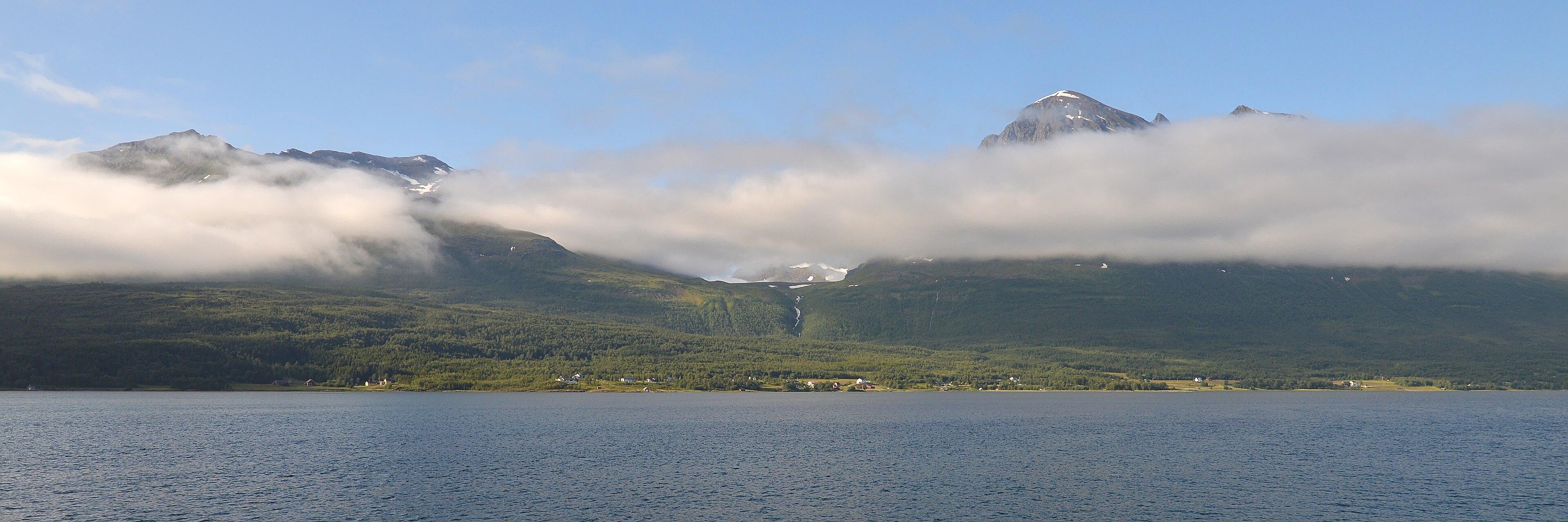 A panoramic photo of a strait with green mountains on it's shore. Low clouds are partly obscuring the mountains. The sky is blue.