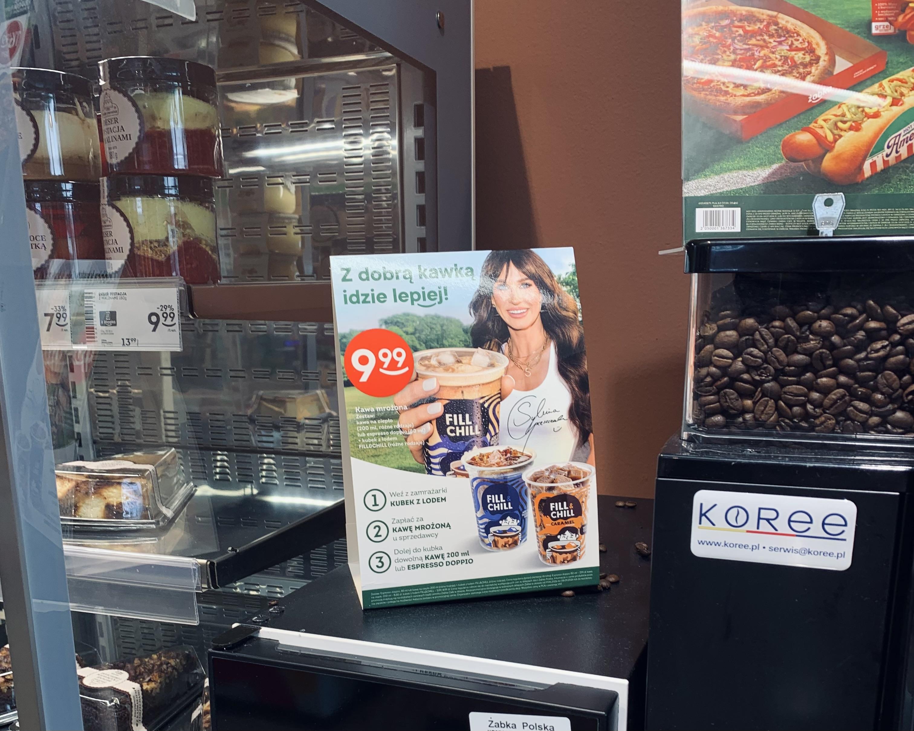 Reklama kawy przy ekspresie w Żabce, na reklamie widoczna Sylwia Grzeszczak podająca kubek kawy wielką ręką.