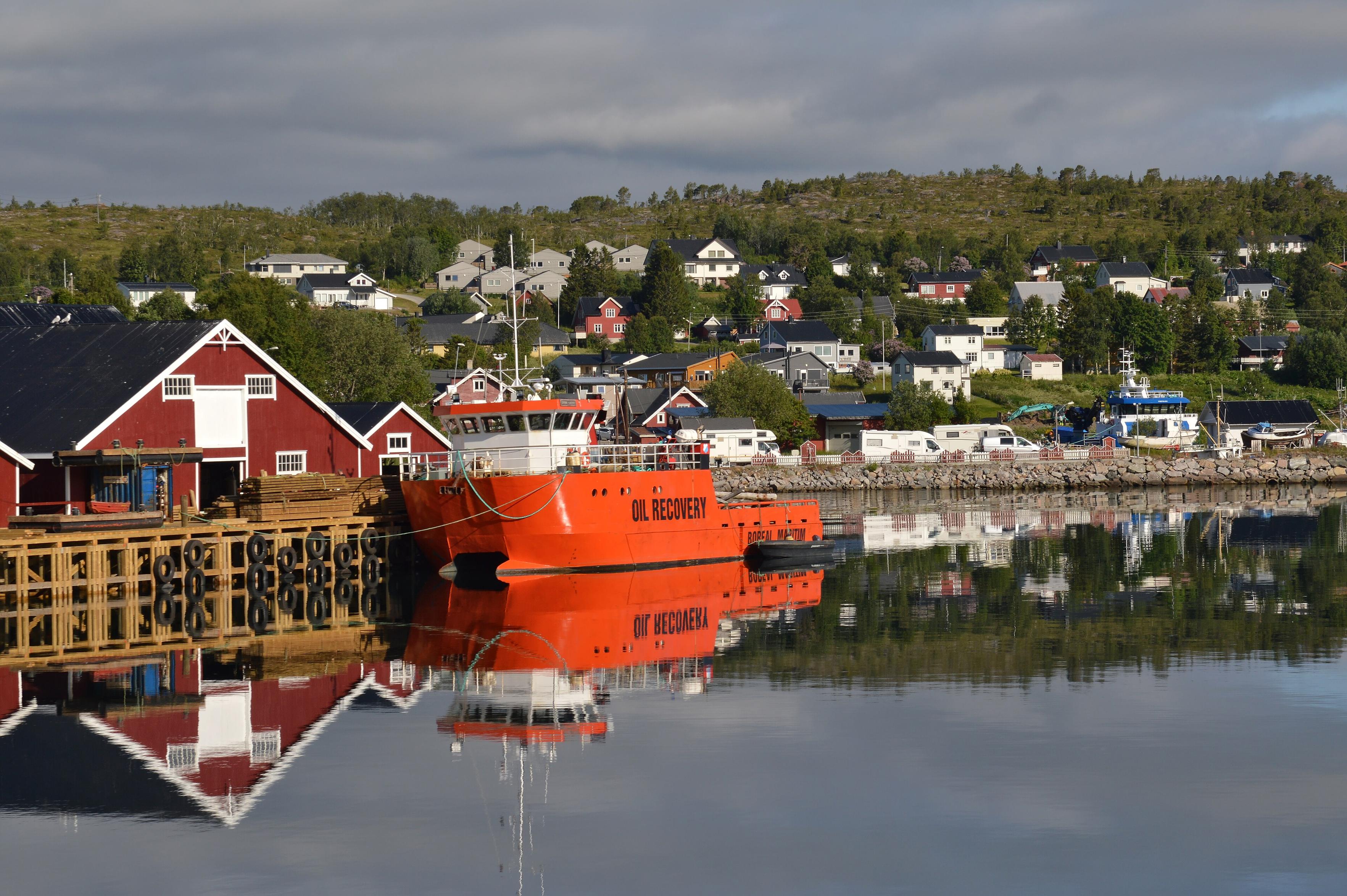 A photo of a village going up a hill on the shore of a strait. There is an orange and white ship with "Oil Recovery" written on it's side in black. The sky is filled with clouds.