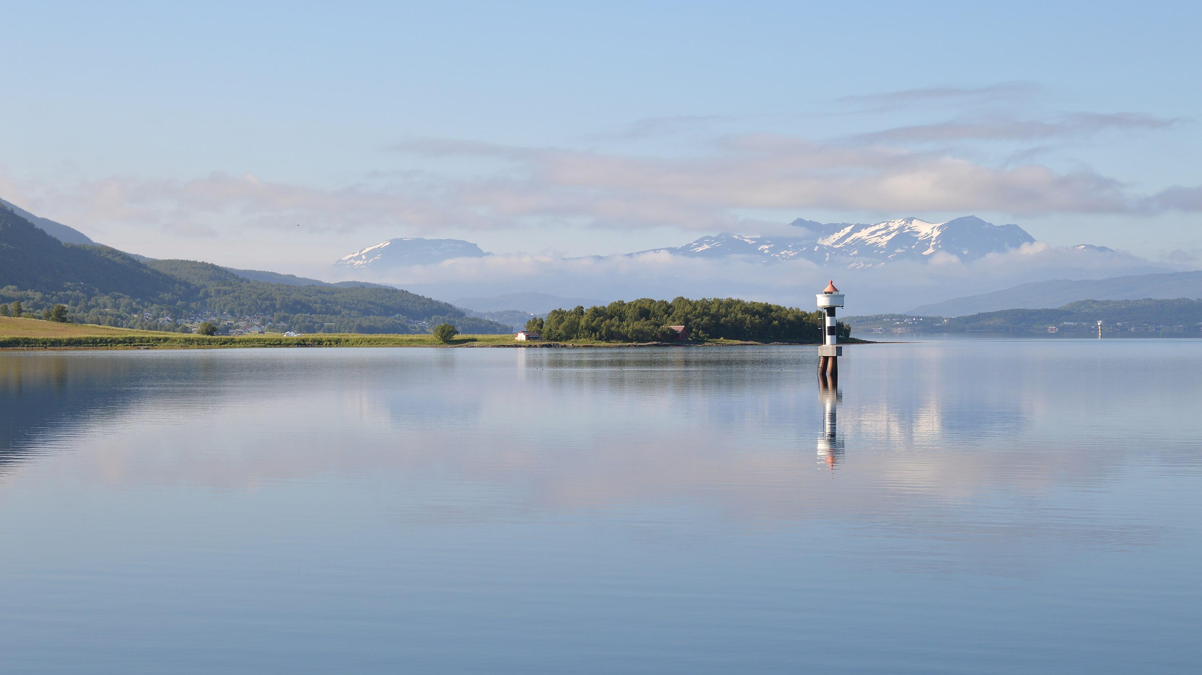 A photo of a strait with mountains in the distance. There is some snow on the mountains and a navigational beacon in the strait. There's a patch of forest in the center of the photo and going up the mountains on the left side. The sky is clear and blue with low clouds moving around the distant mountains.
