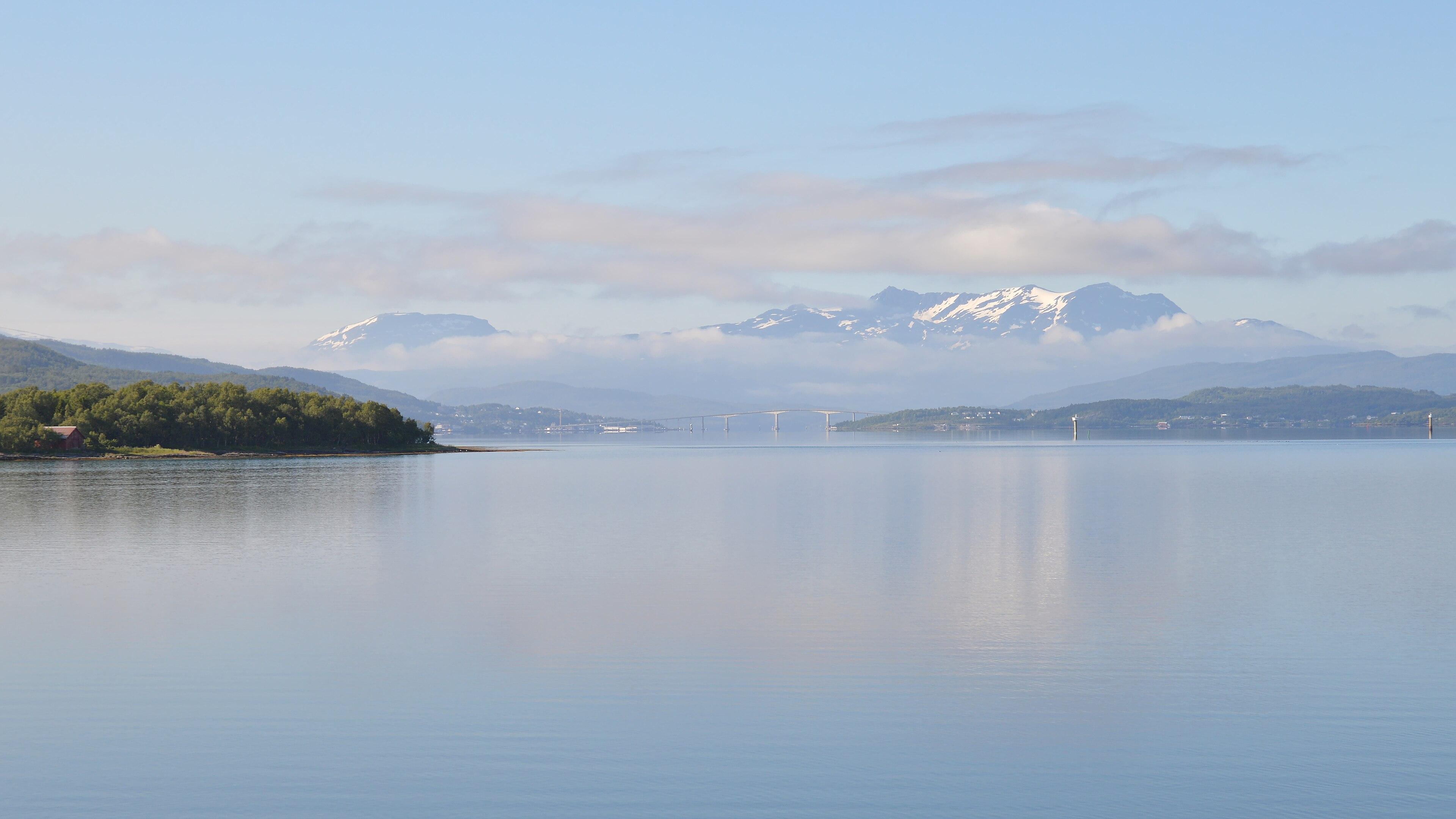 A photo of a strait with a bridge and mountains in the distance. There is some snow on the mountains. The sky is clear and blue with low clouds moving around the distant mountains.