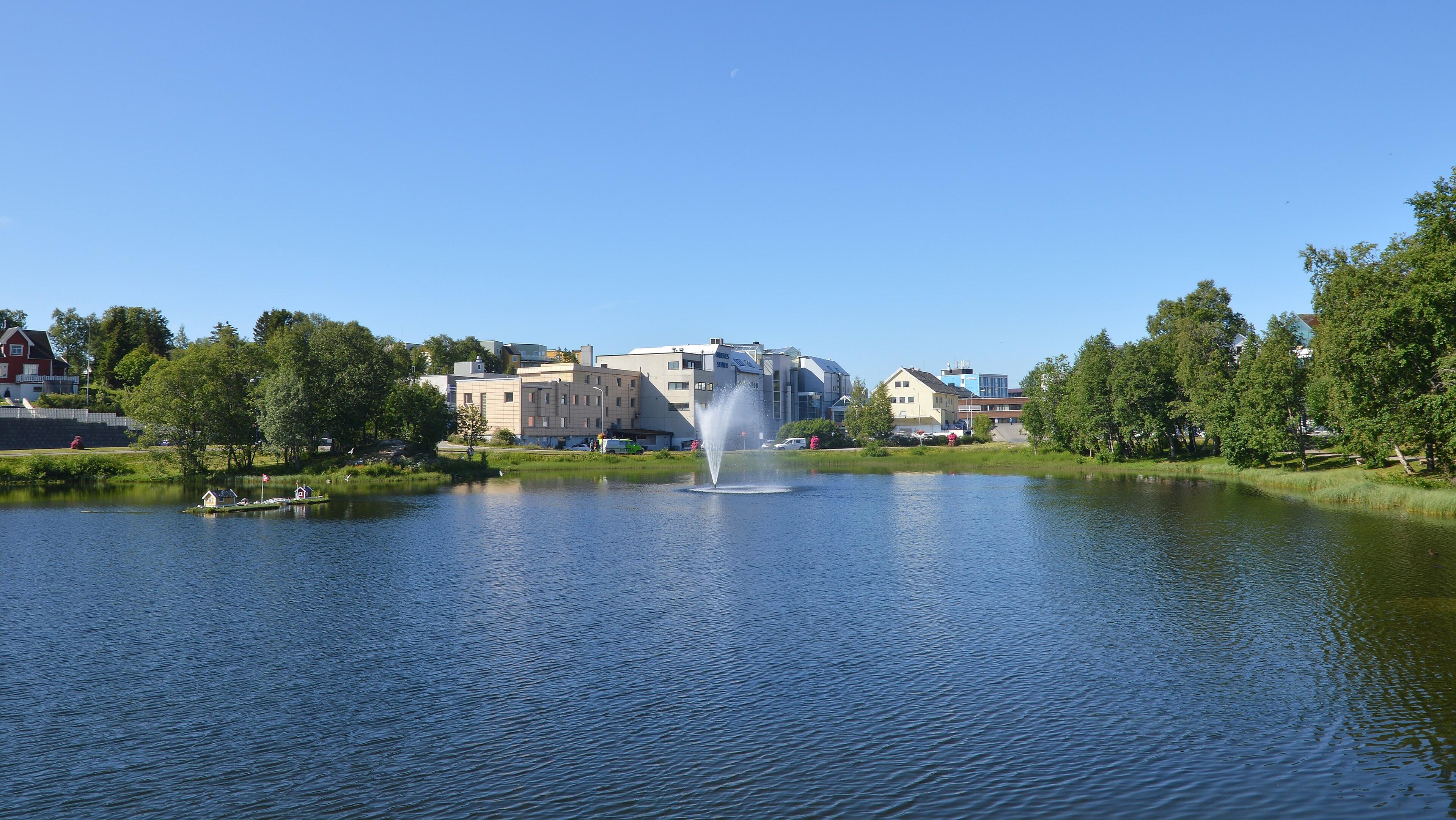 A photo of a small lake with a fountain in it. There are buildings and trees on the far side of the lake. The sky is clear and blue.